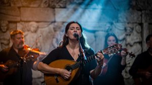 Scène de concert intimiste mêlant mythologie grecque et musiques actuelles, avec une femme chantant et une mandoline, sous un éclairage théâtral chaud et bleu, ambiance feutrée et brumeuse.